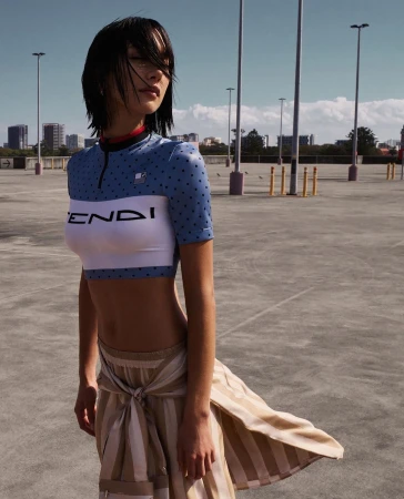 Young woman in blue and white crop top and striped skirt posing in outdoor parking lot setting on sunny day with blue sky and city view.