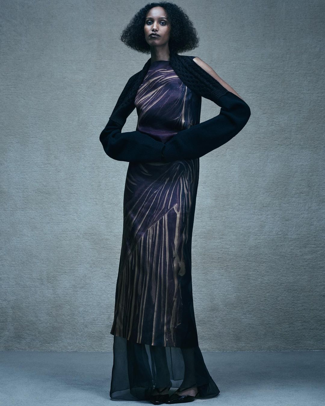 A full-length shot showcases a Black female model in a striking patterned maxi dress and unique knitted arm sleeves, set against a grey backdrop.