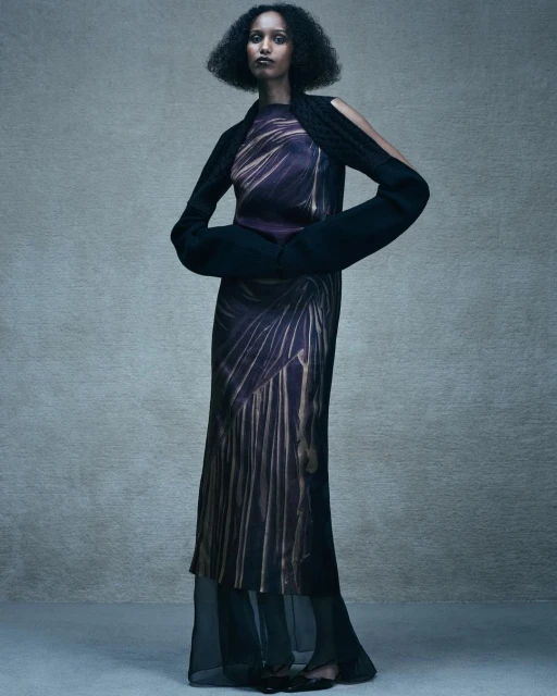 A full-length shot showcases a Black female model in a striking patterned maxi dress and unique knitted arm sleeves, set against a grey backdrop.