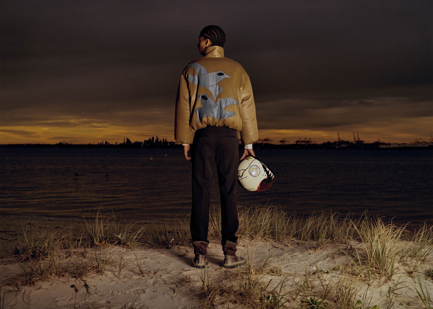 Man stands at dusk on a beach in a brown jacket with a helmet. Water, sand, and city lights are in the background.