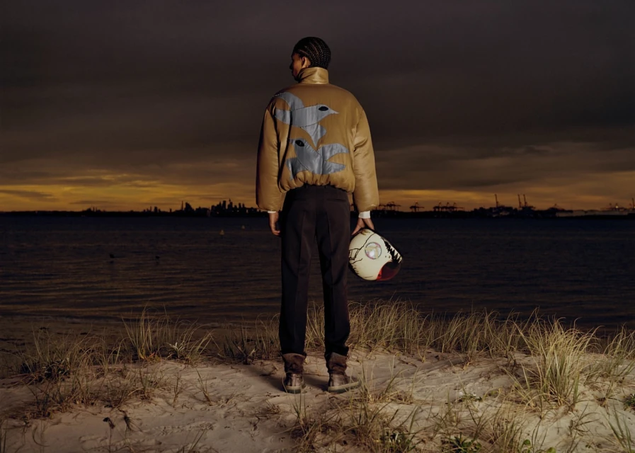 Man stands at dusk on a beach in a brown jacket with a helmet. Water, sand, and city lights are in the background.