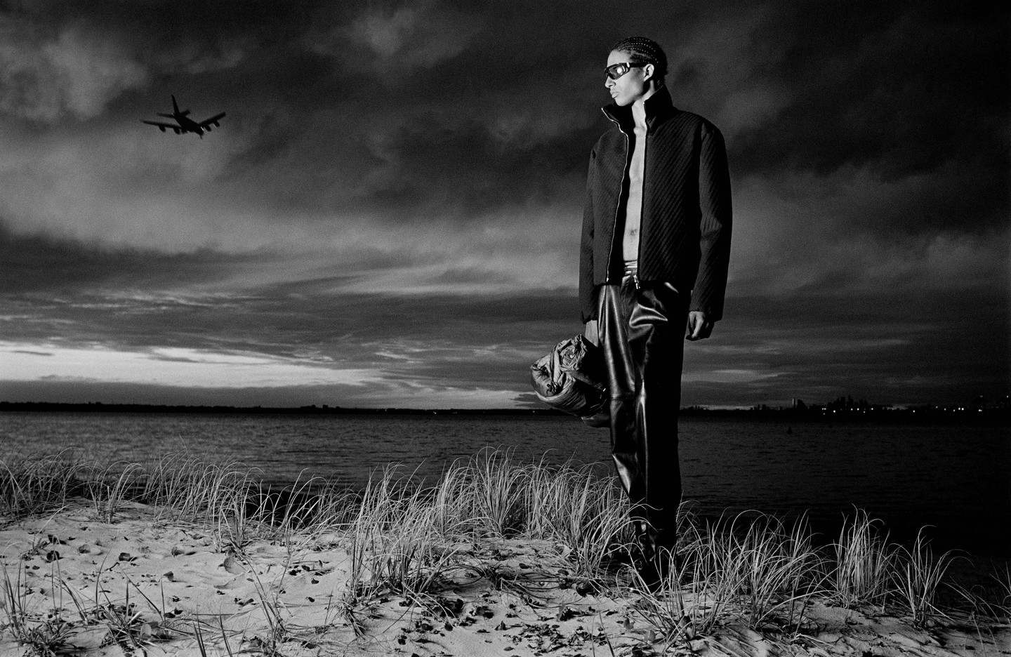 Black and white fashion shot of a man in a jacket and leather pants standing on a beach with an airplane in the cloudy sky.