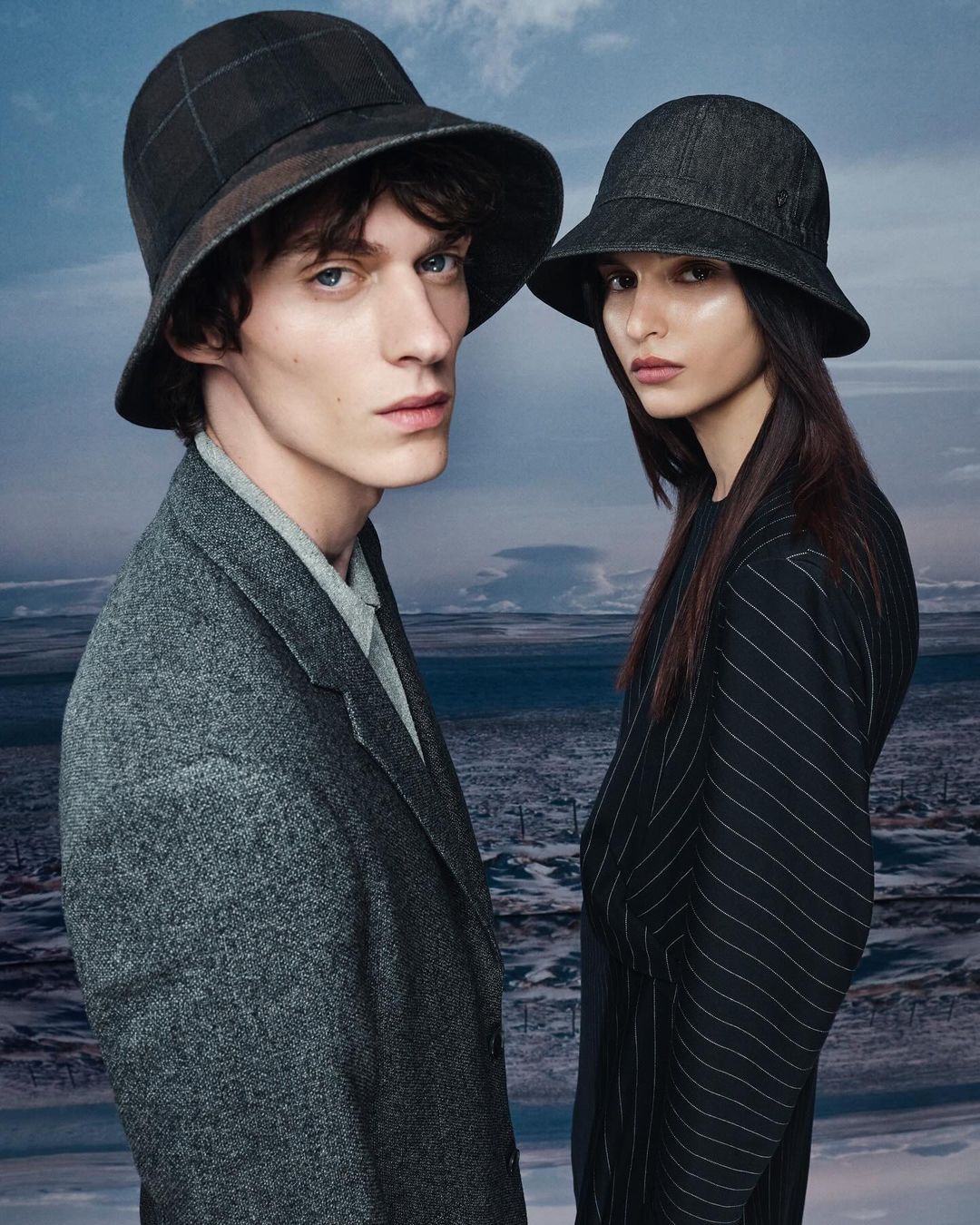 Two fashion models, a man and a woman, stand side-by-side in a photo studio wearing matching hats and tailored clothing, against a scenic backdrop.