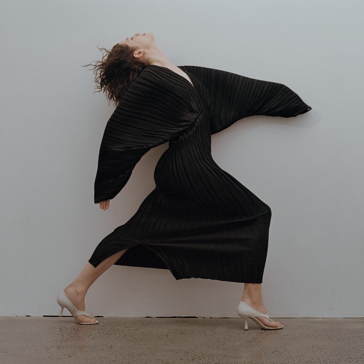 A slender woman with curly red hair dances in a pleated black dress with flowing sleeves against a neutral studio backdrop.