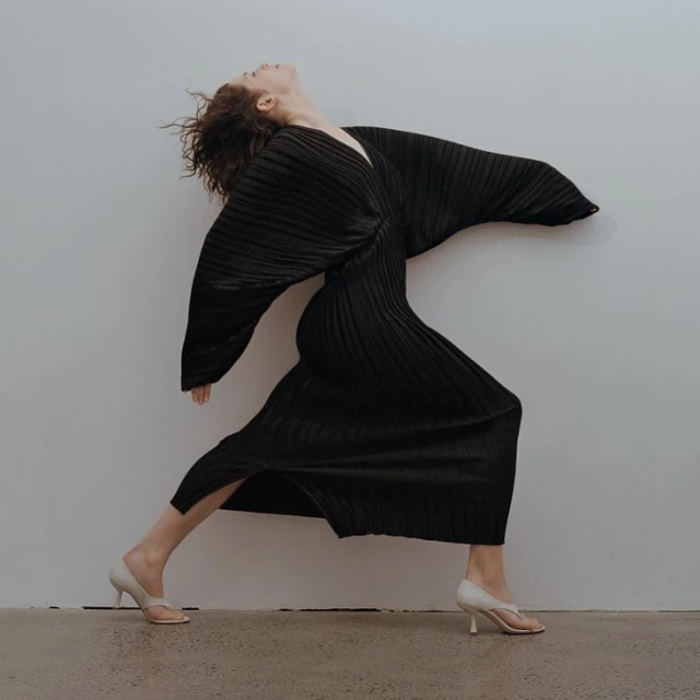 A slender woman with curly red hair dances in a pleated black dress with flowing sleeves against a neutral studio backdrop.