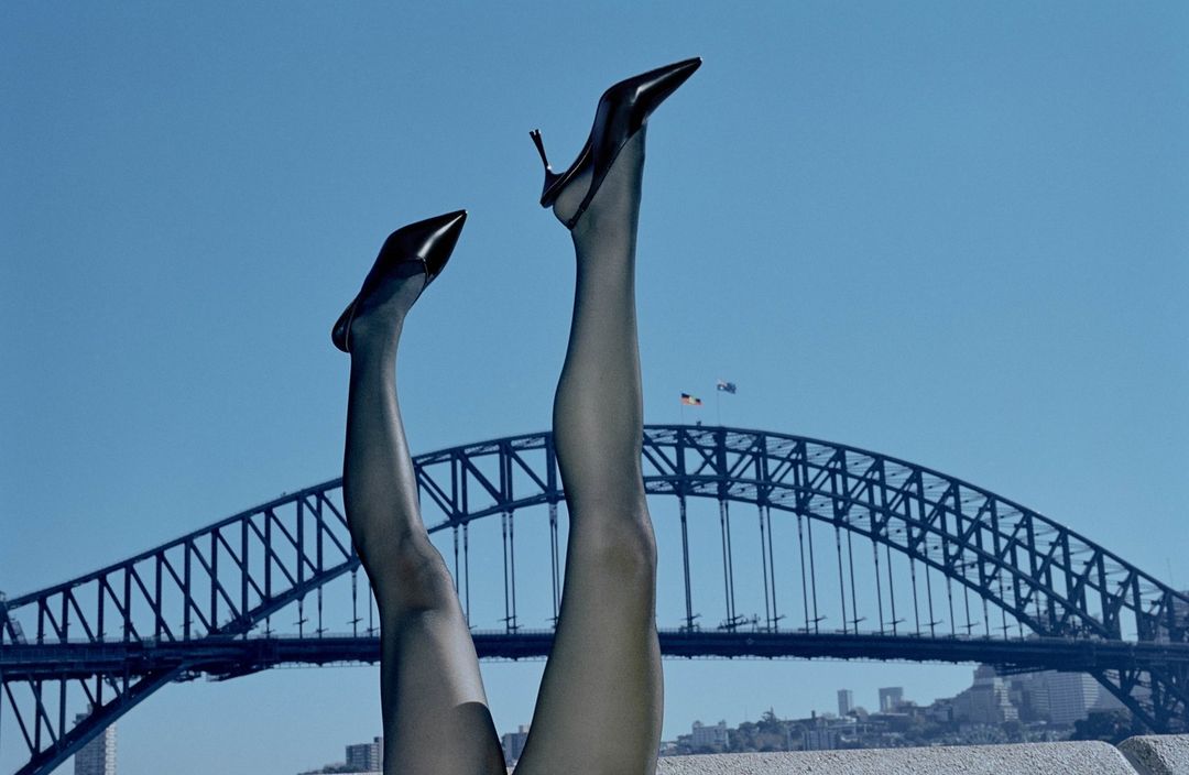 Woman's legs in black stockings and heels point toward a clear blue sky with the Sydney Harbour Bridge in the distance, creating a fashion statement.
