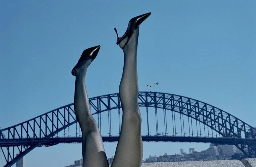 Woman's legs in black stockings and heels point toward a clear blue sky with the Sydney Harbour Bridge in the distance, creating a fashion statement.