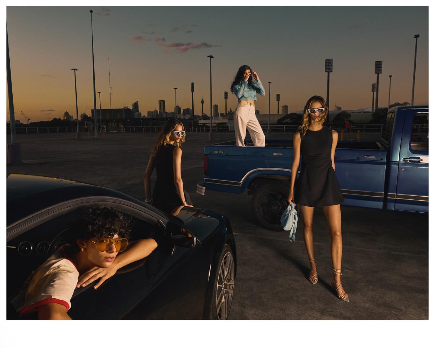 Three women pose with a black car and blue pickup truck against a sunset cityscape backdrop in an urban parking lot.
