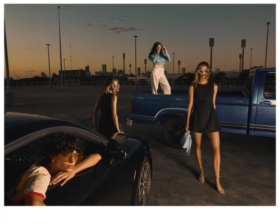 Three women pose with a black car and blue pickup truck against a sunset cityscape backdrop in an urban parking lot.