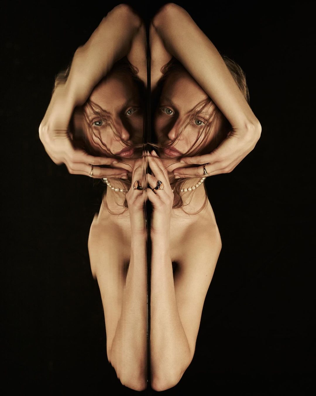 Woman's mirrored portrait with hands framing face, nude shoulders, jewelry, and dark background in studio shot with soft, warm lighting.