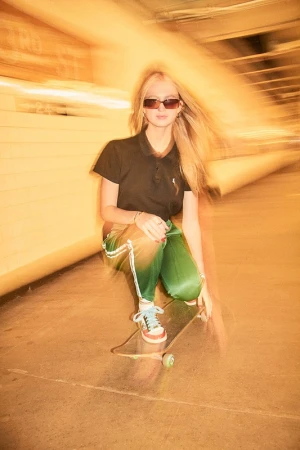 Blonde woman in dark sunglasses and black polo squatting on a skateboard with green pants in a tunnel.