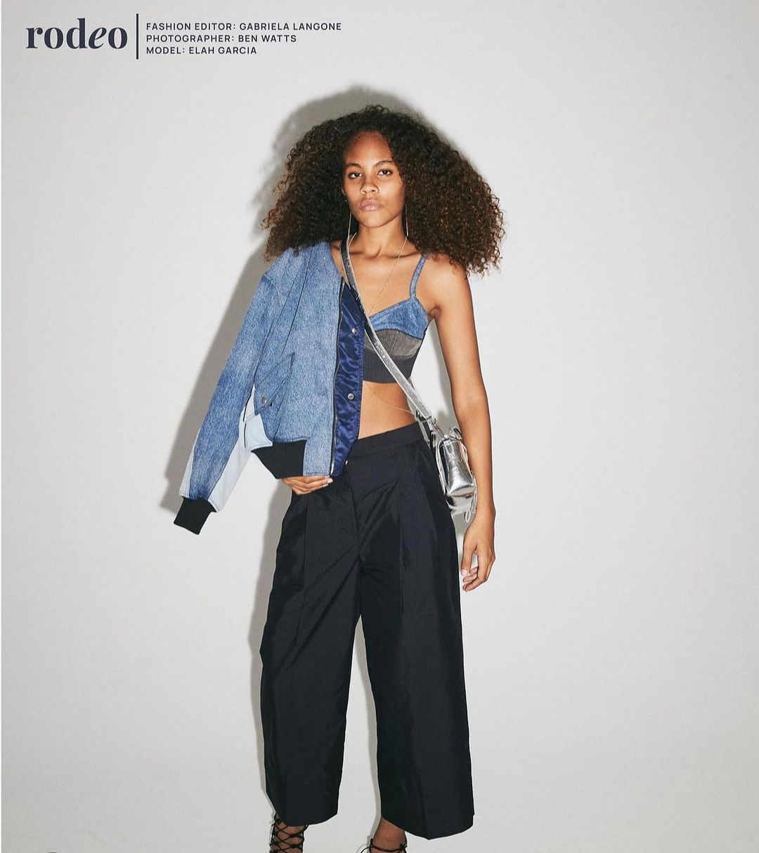 Fashion editorial shot of a young woman with curly hair wearing a blue denim jacket, crop top, and wide-leg pants, posing indoors.