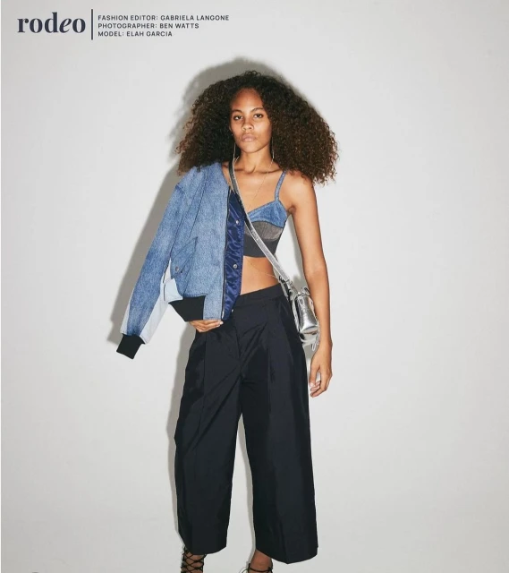 Fashion editorial shot of a young woman with curly hair wearing a blue denim jacket, crop top, and wide-leg pants, posing indoors.