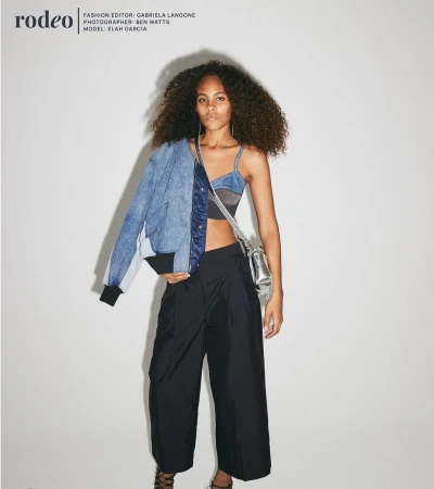 Fashion editorial shot of a young woman with curly hair wearing a blue denim jacket, crop top, and wide-leg pants, posing indoors.