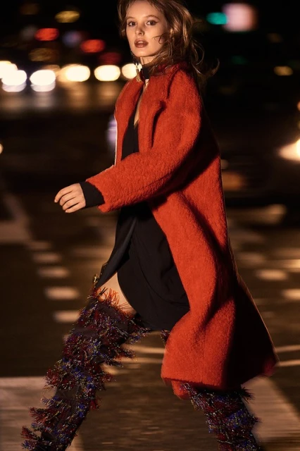A woman in an orange coat and fringed boots walks on a city street at night, with blurred lights creating a vibrant background.