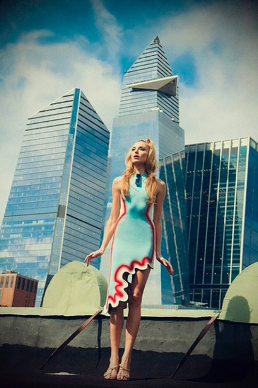 Model wearing a light blue dress with red trim standing on a rooftop against a backdrop of modern glass skyscrapers.
