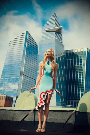 Model wearing a light blue dress with red trim standing on a rooftop against a backdrop of modern glass skyscrapers.