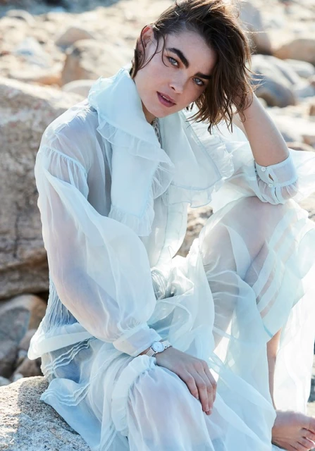 A young woman in a light blue, semi-sheer dress sits on rocks in an outdoor setting with a contemplative expression. Fashion shot.