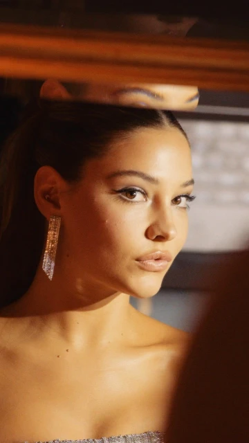 Close-up portrait of a young woman reflecting in a mirror with visible makeup, earring, and bare shoulder in warm, studio lighting.