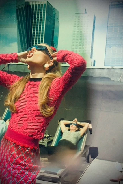 Two women wearing sunglasses and posed against a city building backdrop on a bright day. The woman in the foreground is wearing red.