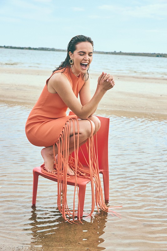 Smiling woman in orange fringed dress squatting on a red plastic chair standing in shallow water