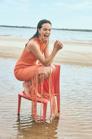 Smiling woman in orange fringed dress squatting on a red plastic chair standing in shallow water