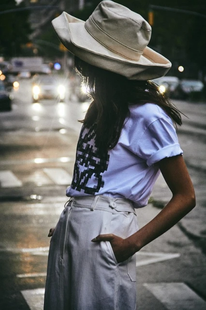 A woman in a hat stands on a crosswalk in a city, wearing a white t-shirt and pants in an urban setting with cars and streetlights visible.