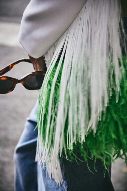 Fashionable woman wearing a white blazer and blue jeans, accessorized with sunglasses and a white-and-green fringe purse.