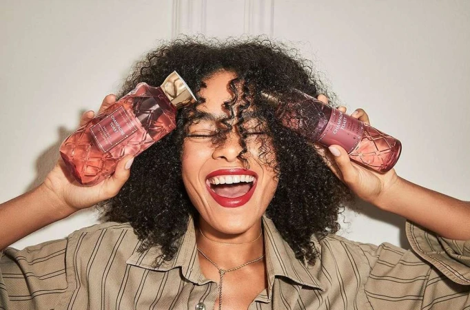Cheerful woman with curly hair smiles widely holding two bottles of champagne toast body wash against a cream-colored wall.