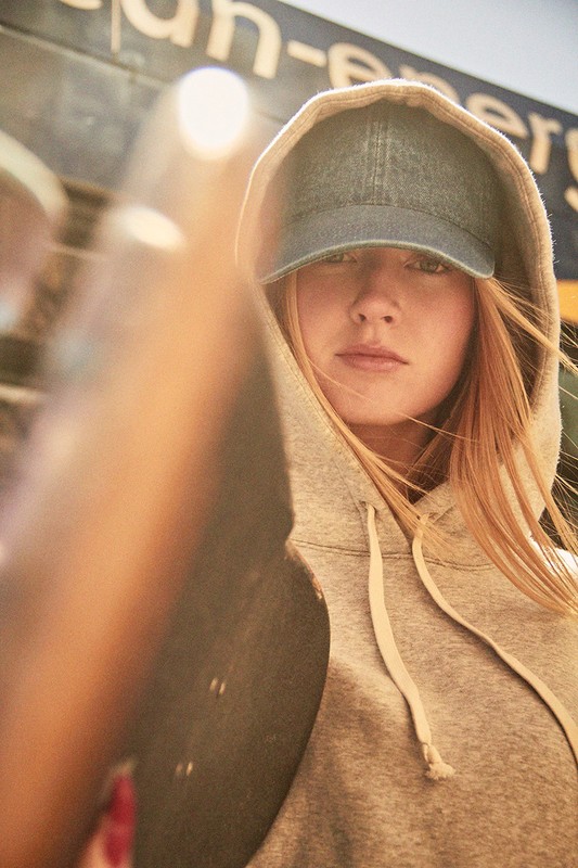 Portrait of an adult woman in a hoodie and baseball cap holding a skateboard, posing in an urban setting with diffused light.