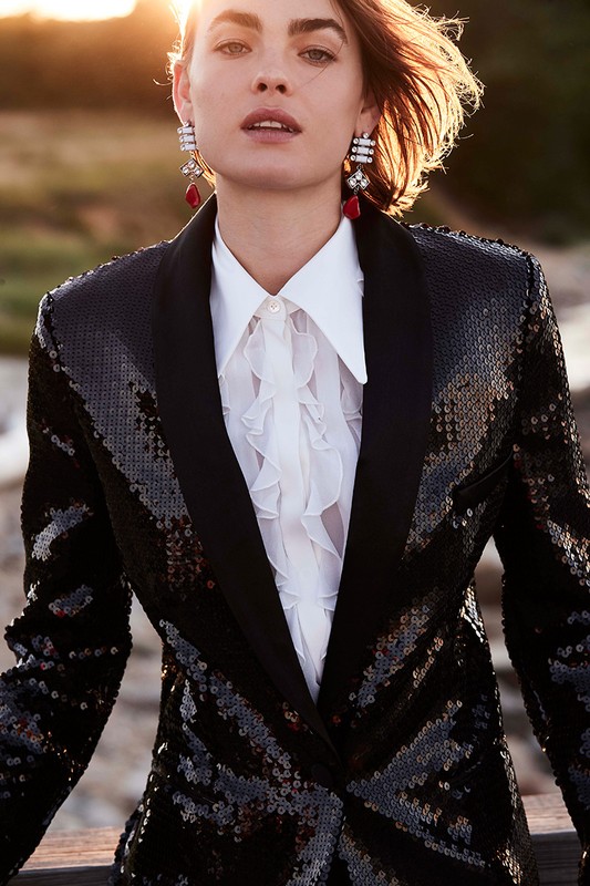 Fashion shot of a woman wearing a sequined blazer, white ruffled shirt, and statement earrings posing against a warm, natural backdrop.