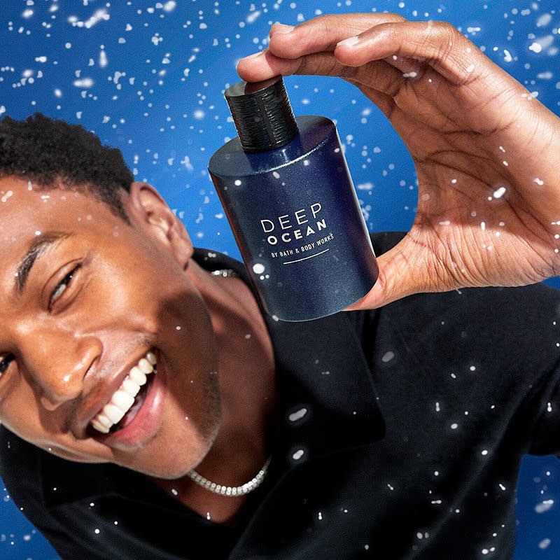 Smiling African-American man holds and presents a blue bottle of Deep Ocean by Bath & Body Works against a blue background with snow.