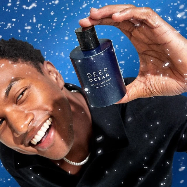 Smiling African-American man holds and presents a blue bottle of Deep Ocean by Bath & Body Works against a blue background with snow.