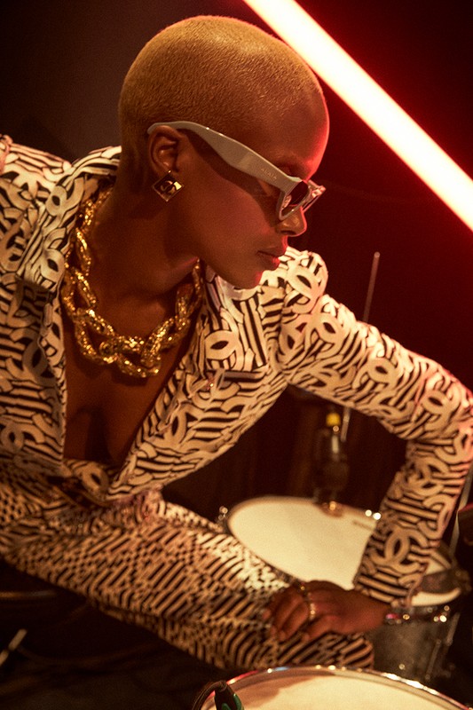 Close-up studio shot of a Black woman in patterned suit with gold jewelry playing the drums, illuminated by red neon lights.