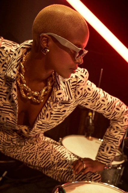 Close-up studio shot of a Black woman in patterned suit with gold jewelry playing the drums, illuminated by red neon lights.