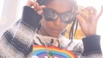 Black woman wearing cat-eye sunglasses and a patterned sweater holding her glasses frame by Ben Watts