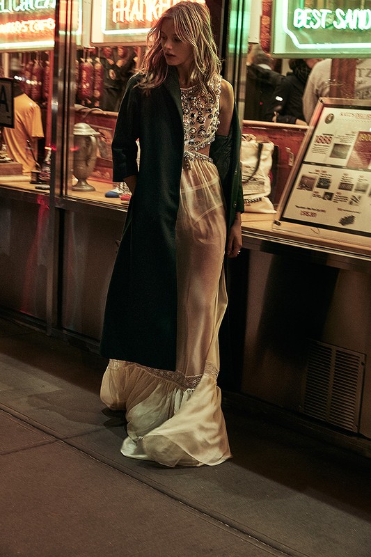 A fashion model in a shimmering top and dress walks past a diner at night in an outdoor urban setting with illuminated signs.
