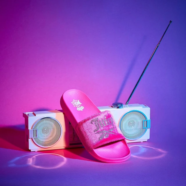 Pink slide sandal displayed with a vintage-style boombox against a purple and pink gradient backdrop in a studio product shot.