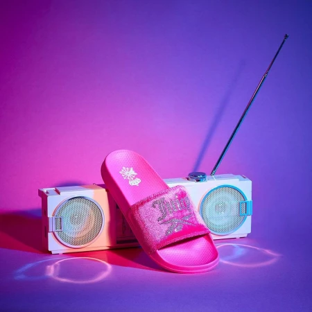 Pink slide sandal displayed with a vintage-style boombox against a purple and pink gradient backdrop in a studio product shot.