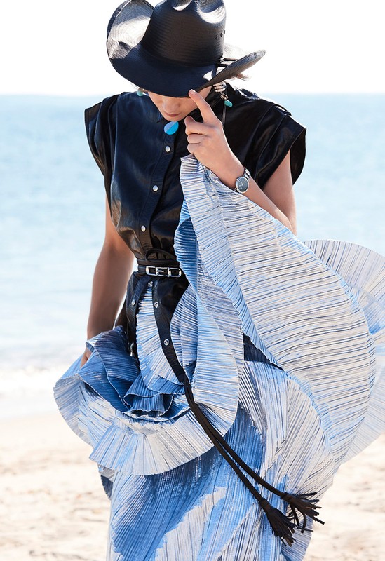 Woman in elegant black hat and pleated dress stands on beach with blue water. Fashion shot, eye-level angle, sunny outdoor light.