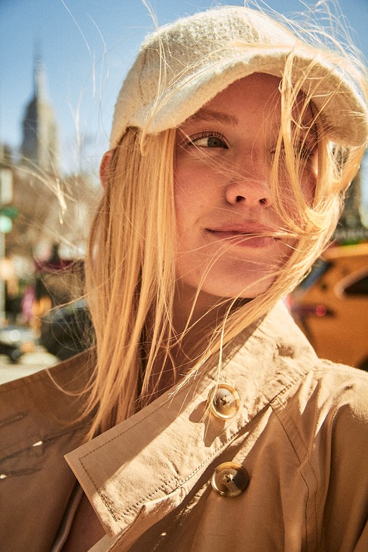 Close-up of a blonde woman wearing a cream cap and tan coat with a city street and skyscraper in the background on a sunny day.