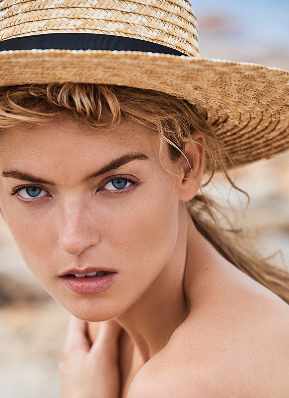Close-up portrait of a young blonde woman wearing a woven sunhat with black ribbon, looking directly at the camera with bright blue eyes.