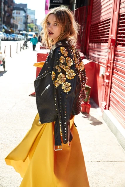 Woman in a black leather jacket with gold floral embellishments and a long yellow skirt standing on a city street.
