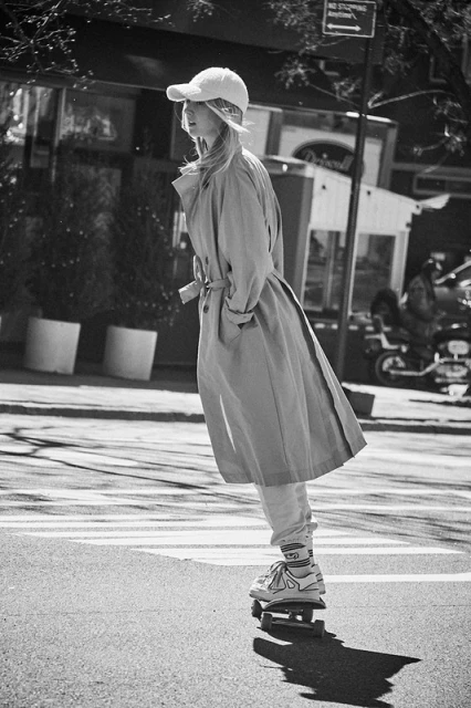Woman wearing a trench coat and baseball cap skateboarding on a city crosswalk in black and white, capturing an urban fashion moment.