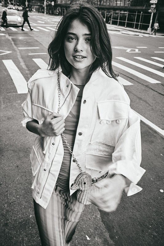 Monochrome portrait of a young woman walking on a city street wearing a utility jacket and holding a small animal-shaped handbag.
