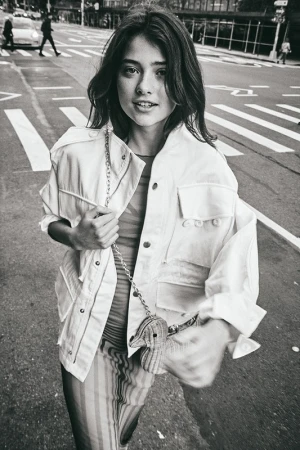 Monochrome portrait of a young woman walking on a city street wearing a utility jacket and holding a small animal-shaped handbag.