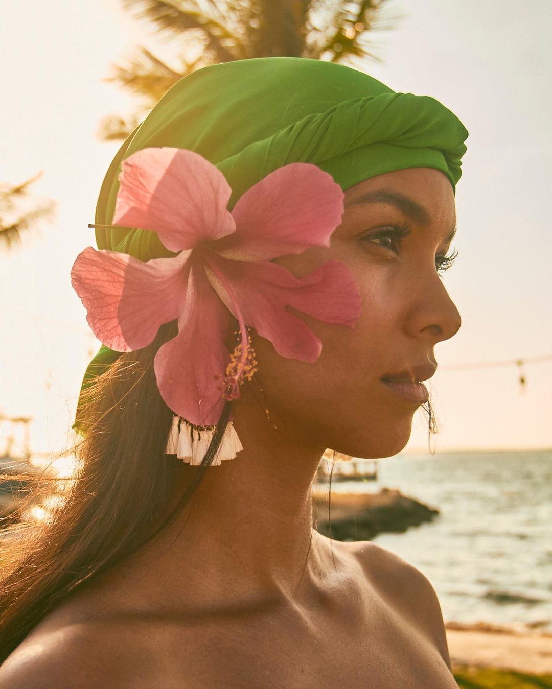 Close-up of woman with green headscarf and pink hibiscus flower by the ocean at sunset. Outdoor portrait with golden light.