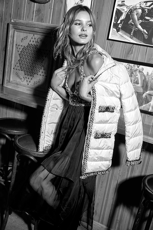 Black and white portrait of a fair-skinned young woman in a sequined dress and puffer jacket posing in a bar setting.