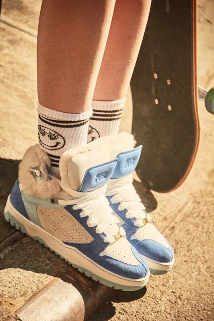 Close-up of fashion sneakers with white fur trim, blue and tan suede, white laces, worn with socks with smiley face graphics.
