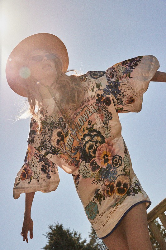 Woman in floral print dress and hat in low-angle shot against blue sky with bright sunlight, capturing a fashion-forward outdoor moment.